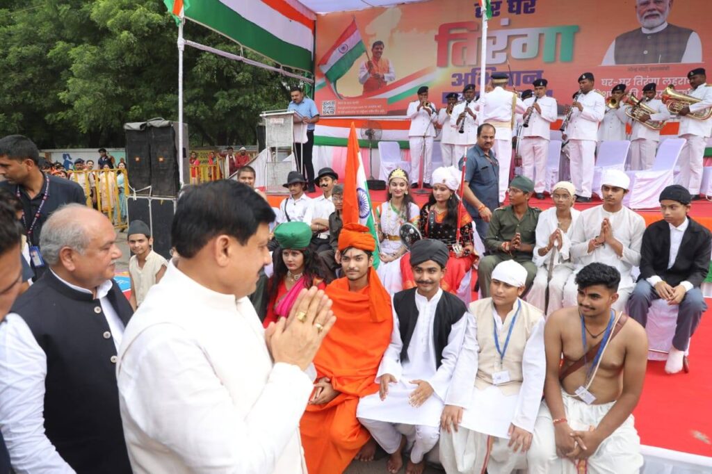 मुख्यमंत्री डॉ. मोहन यादव ग्वालियर में हर घर तिरंगा अभियान के तहत आयोजित कार्यक्रम में महापुरुषों की वेशभूषा में सजे बच्चों का अभिवादन करते हुए, पृष्ठभूमि में बैंड दल और मंच पर तिरंगा अभियान का पोस्टर।