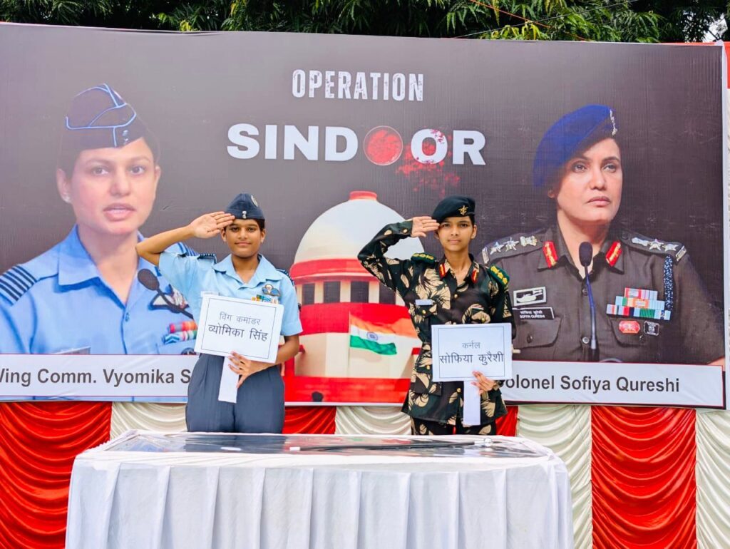 ऑपरेशन सिंदूर कार्यक्रम में भारतीय वायुसेना और सेना की वर्दी में सजे दो युवा, सलामी देते हुए, पृष्ठभूमि में विंग कमांडर व्योमिका सिंह और कर्नल सोफिया कुरैशी के पोस्टर।